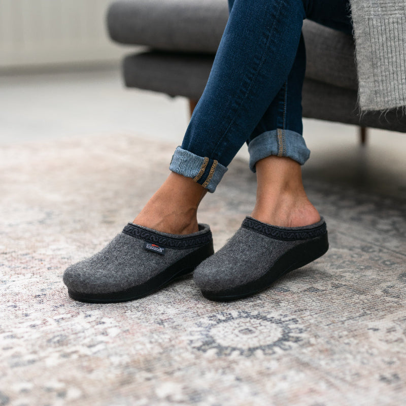 A person sits cross-legged on a couch, wearing rolled-up jeans and Women's WoolFlex Wool Clog - Medium in grey, on a patterned rug.