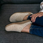 A woman in beige Women's Eco Wool Clog slippers with a cork latex footbed and dark blue jeans relaxes on a gray couch.