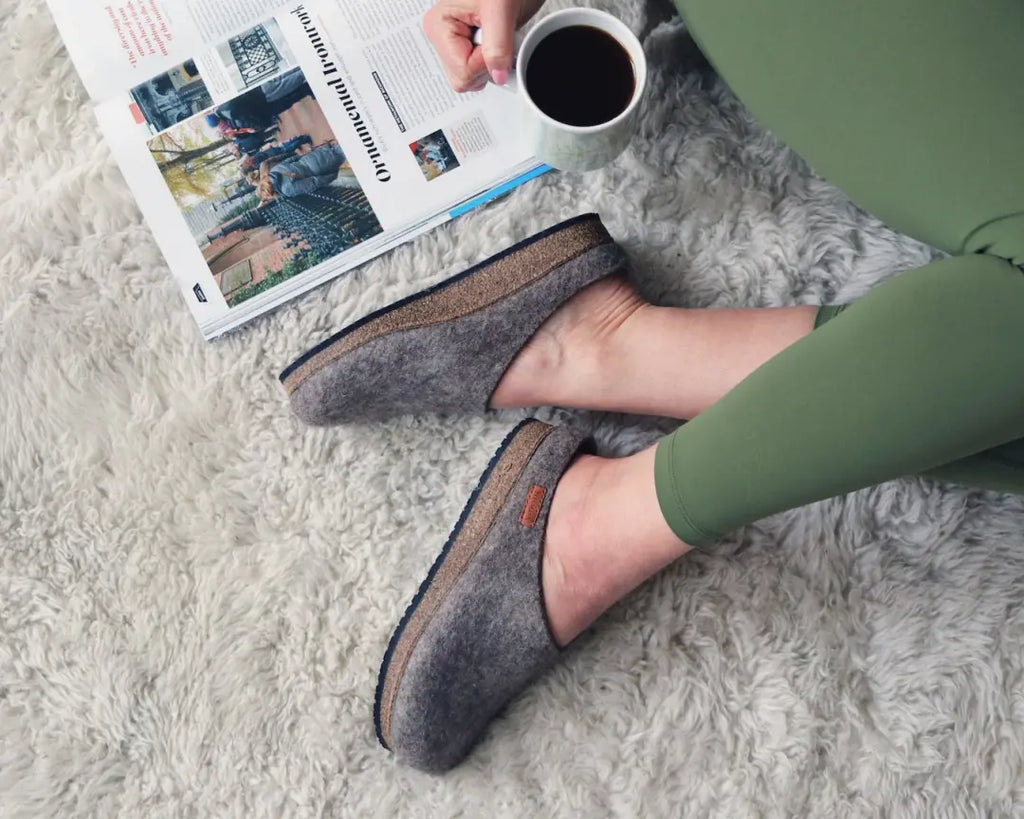A person wearing Women's Eco Wool Clog with a cork latex footbed and green leggings sits on a light rug, sipping coffee and reading a magazine.