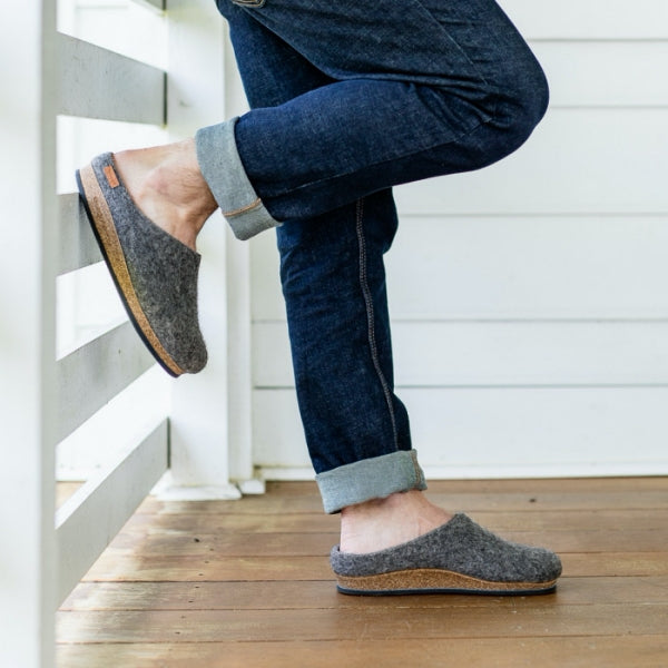 A person in rolled-up jeans leans against a white railing, wearing the Men's Eco Wool Clog—grey felt slip-on slippers crafted from sustainable wool with cork soles—on a wooden porch.
