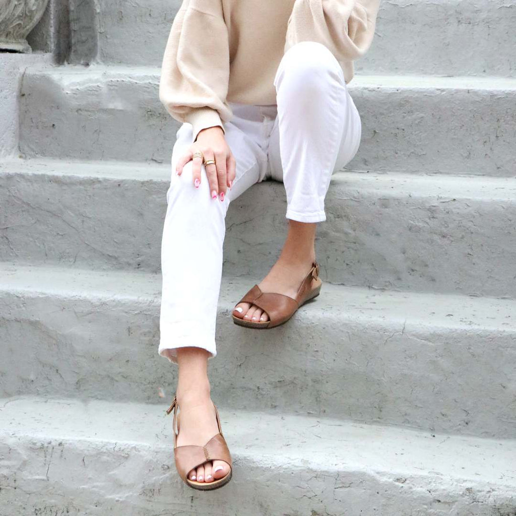 On gray steps, a person sits wearing a beige sweater and white pants paired with Women's Louisa Leather Slingback Sandals in brown, featuring a cushioned footbed. Their hand rests on their knee.
