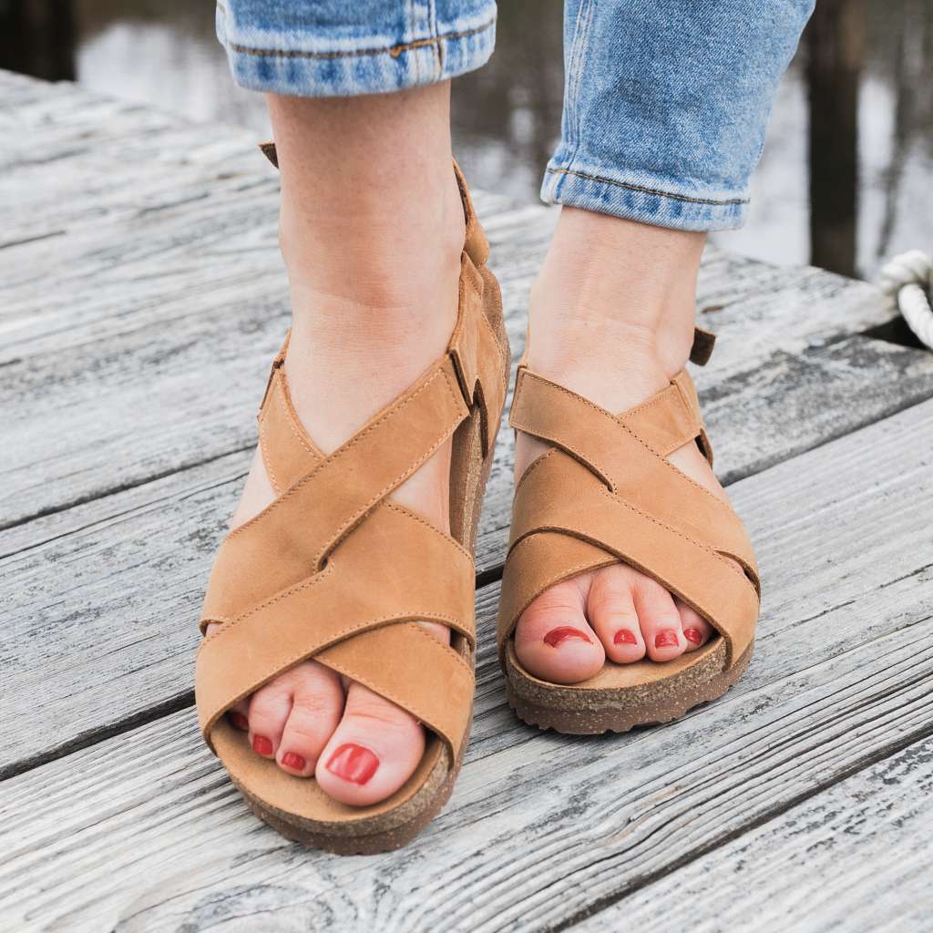 A person wearing the Women's Hannah Leather Sandal with a suede footbed and blue jeans stands on a weathered wooden surface.