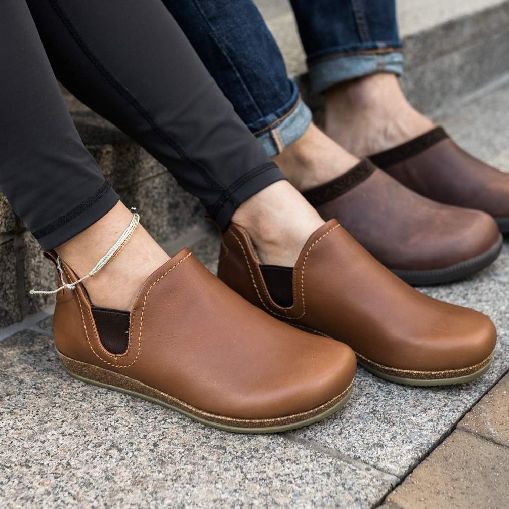 Two people sit on concrete steps, both wearing brown Women's Lieben Leather Chelsea Boots. One wears leggings with an ankle bracelet, while the other sports jeans. The boots feature a soft wool felt lining, adding a cozy touch to their casual style.