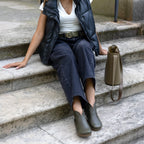 A person sits on stone steps wearing a black vest, white shirt, and dark pants cinched with the Women’s Matching Leather Belt. Green shoes complete the look, and a taupe handbag with a strap rests beside them on the steps.