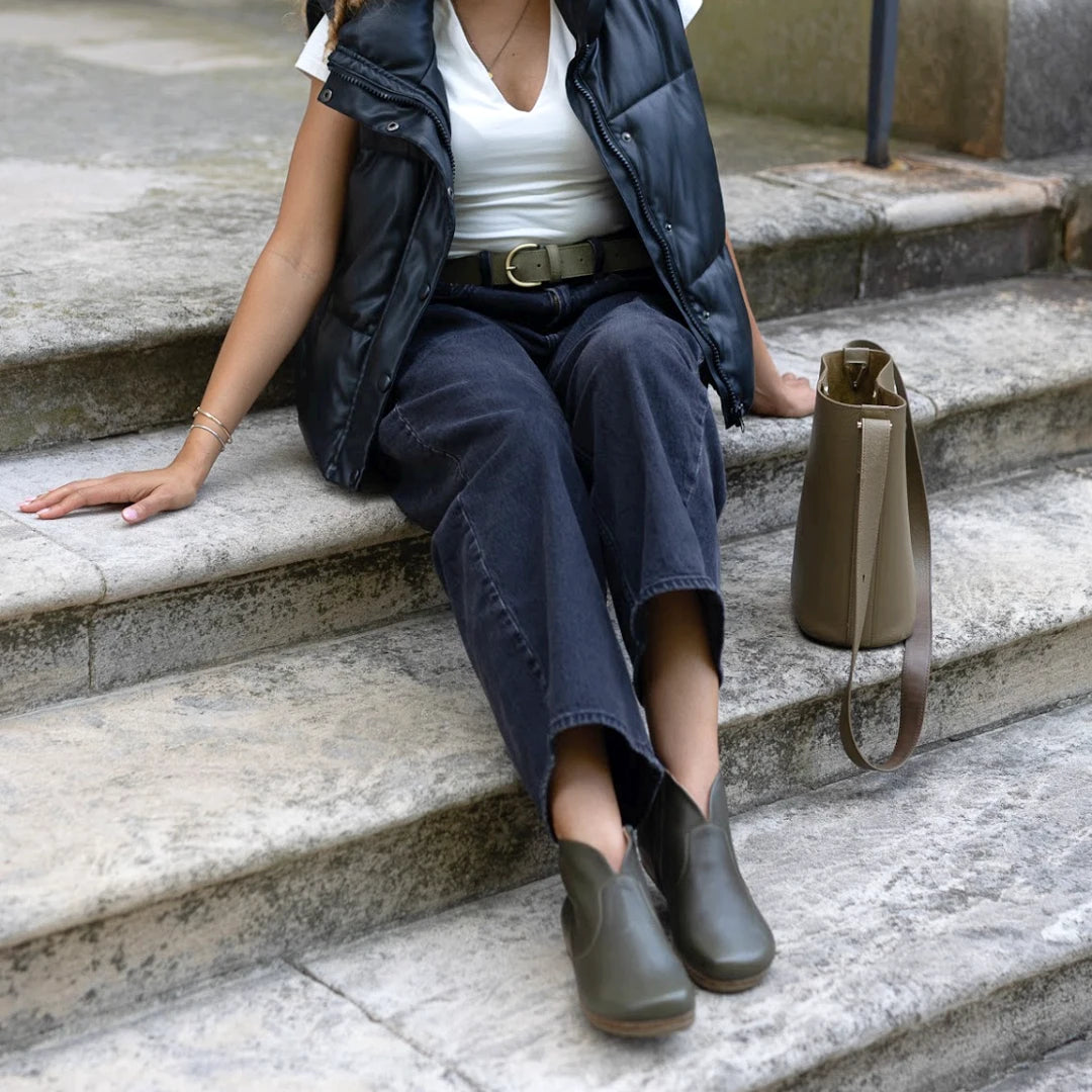 A person sits on stone steps wearing a black vest, white shirt, and dark pants cinched with the Women’s Matching Leather Belt. Green shoes complete the look, and a taupe handbag with a strap rests beside them on the steps.
