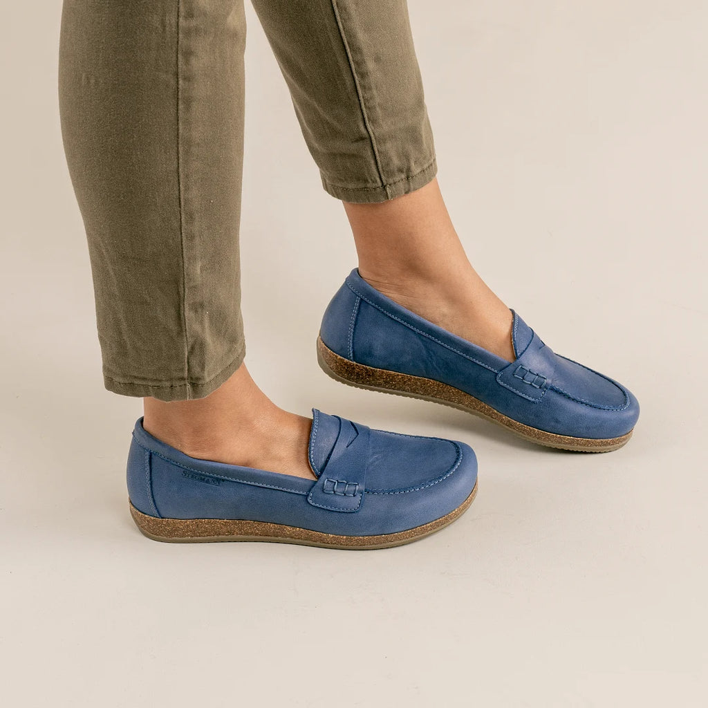A person wearing olive green pants and Women's Victoria Loafer in blue nubuck with cushioned insoles stands on a light-colored floor.