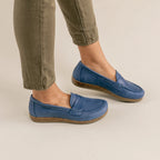 A person wearing olive green pants and Women's Victoria Loafer in blue nubuck with cushioned insoles stands on a light-colored floor.
