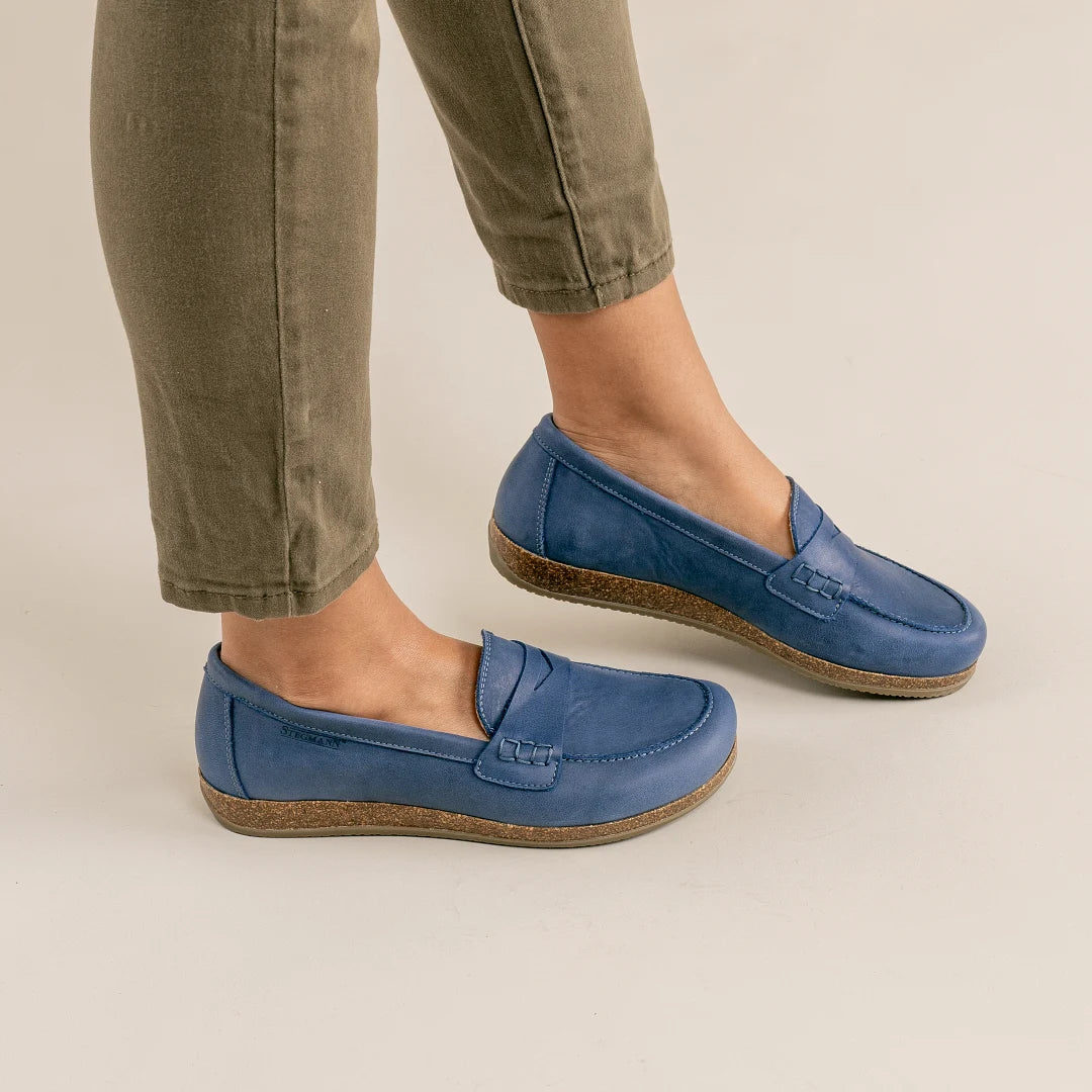 A person wearing olive green pants and Women's Victoria Loafer in blue nubuck with cushioned insoles stands on a light-colored floor.
