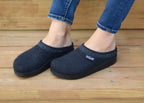 A person stands on wooden steps, wearing dark WoolFlex Wool Clogs with patterned trim and jeans. This stylish footwear provides arch support, ensuring all-day comfort.