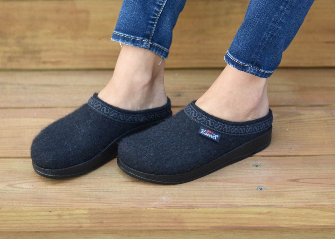 A person stands on wooden steps, wearing dark WoolFlex Wool Clogs with patterned trim and jeans. This stylish footwear provides arch support, ensuring all-day comfort.