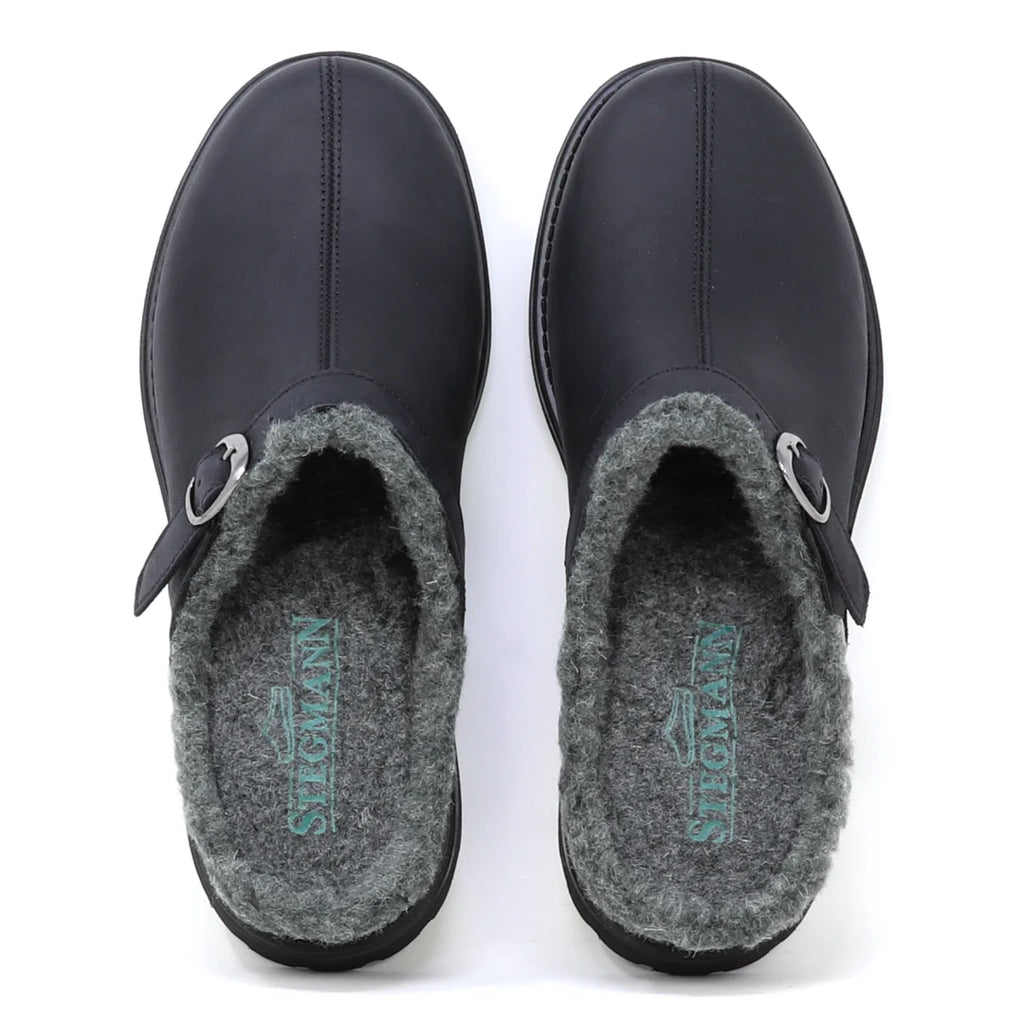 The Zermatt Wool Shearling Clog is shown from above—a pair of black slip-on clogs featuring a wool shearling lining and buckle detail on the left shoe.