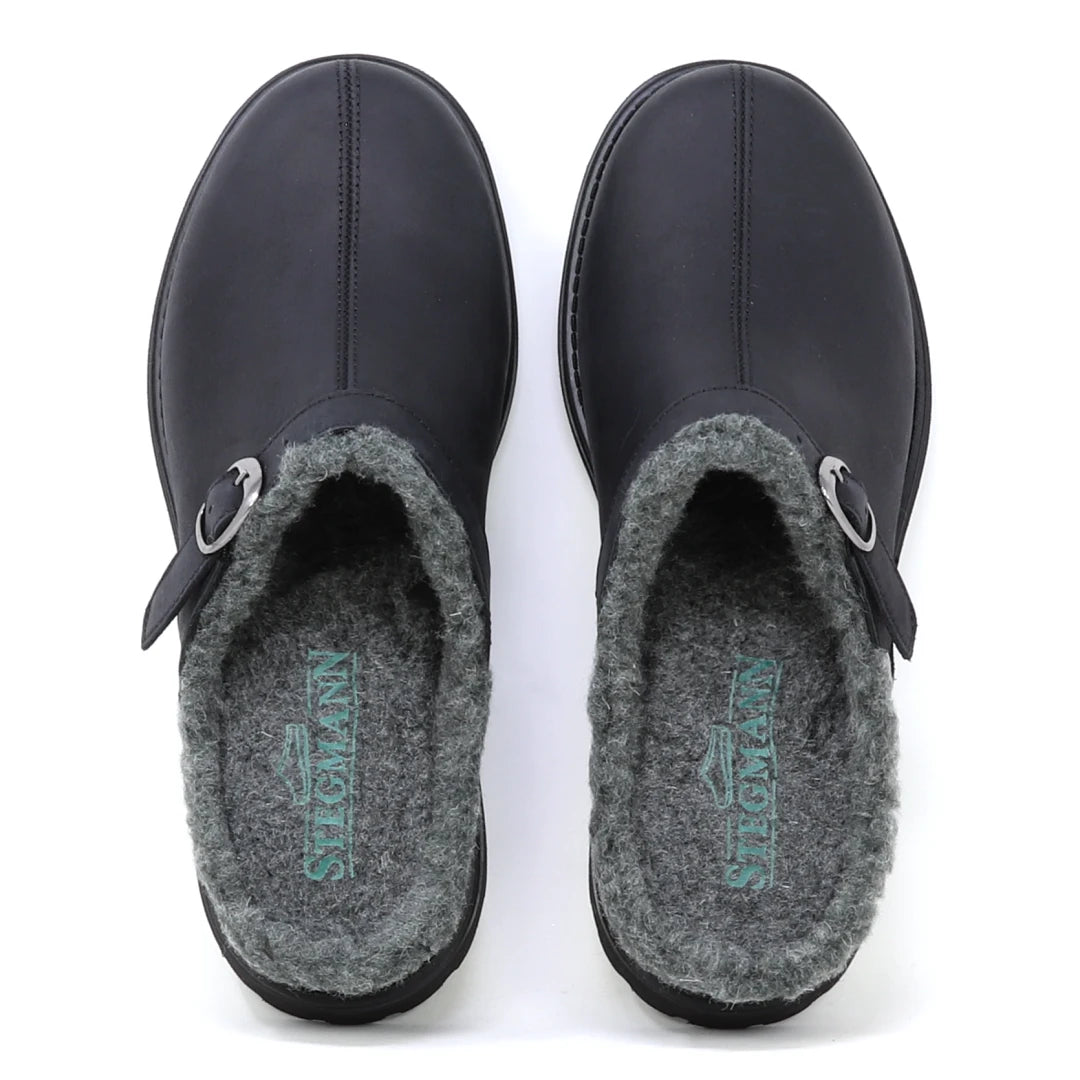 The Zermatt Wool Shearling Clog is shown from above—a pair of black slip-on clogs featuring a wool shearling lining and buckle detail on the left shoe.