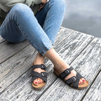 A person in blue jeans and Women's Heidi Leather Sandal with adjustable straps sits on a wooden dock near the water, feet crossed at the ankles.