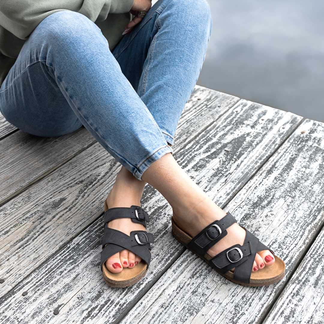 A person in blue jeans and Women's Heidi Leather Sandal with adjustable straps sits on a wooden dock near the water, feet crossed at the ankles.