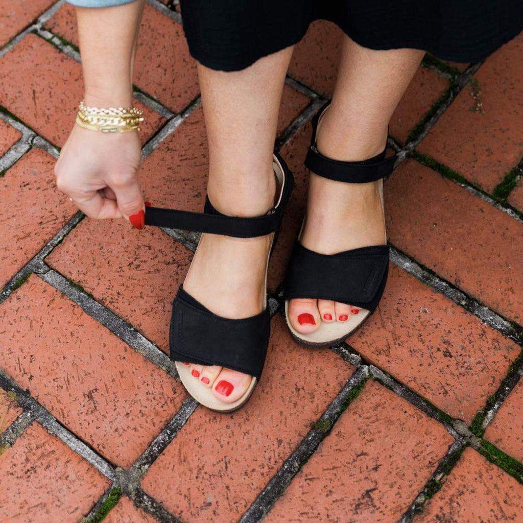 A person fastens the strap of the Women's Freya Leather Wedge Sandal on their foot while standing on a brick pavement.