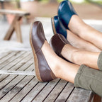 Two people wearing Women’s Liesl Leather Skimmer shoes—one in brown, one in blue, both with anatomically shaped support soles—rest their feet on a wooden slatted bench outdoors.