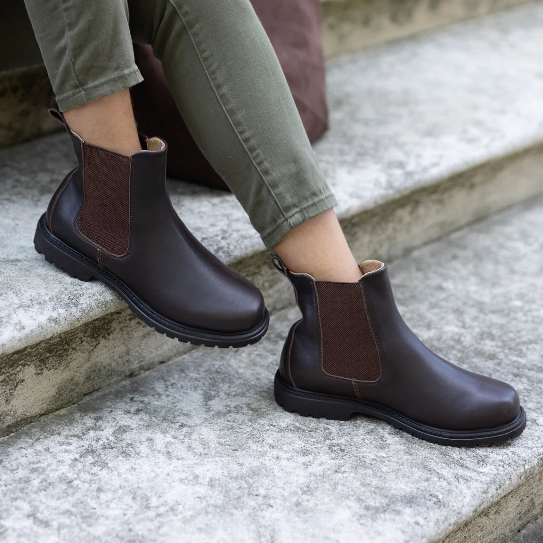 A person wearing the Women’s Brenner Chelsea Boot in dark brown water-resistant leather with olive green pants, sitting on outdoor concrete steps.