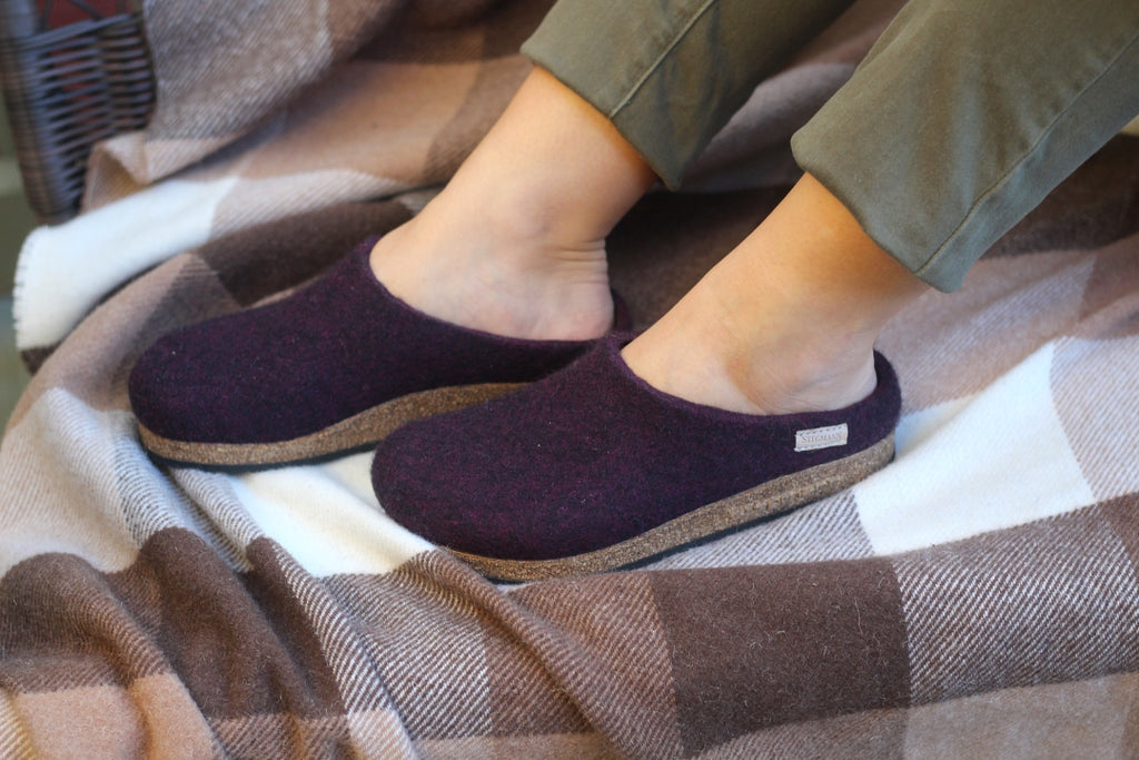 A person wearing Women's Essenz Wool Clog slippers in dark purple with cork soles sits on a brown and white plaid blanket; only their lower legs and feet are visible.