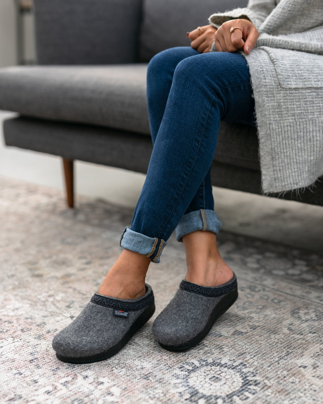 A person sits on a sofa wearing blue jeans, a gray sweater, and the Women's Original 108 Wool Clog - Medium with an orthopedically shaped sole, legs crossed and hands resting on their knee.