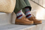 A person in green pants, colorful patterned socks, and Women's Mainz Leather Moc Shoe with an UltraComfort footbed sits on the stairs, a woven bag by their side.