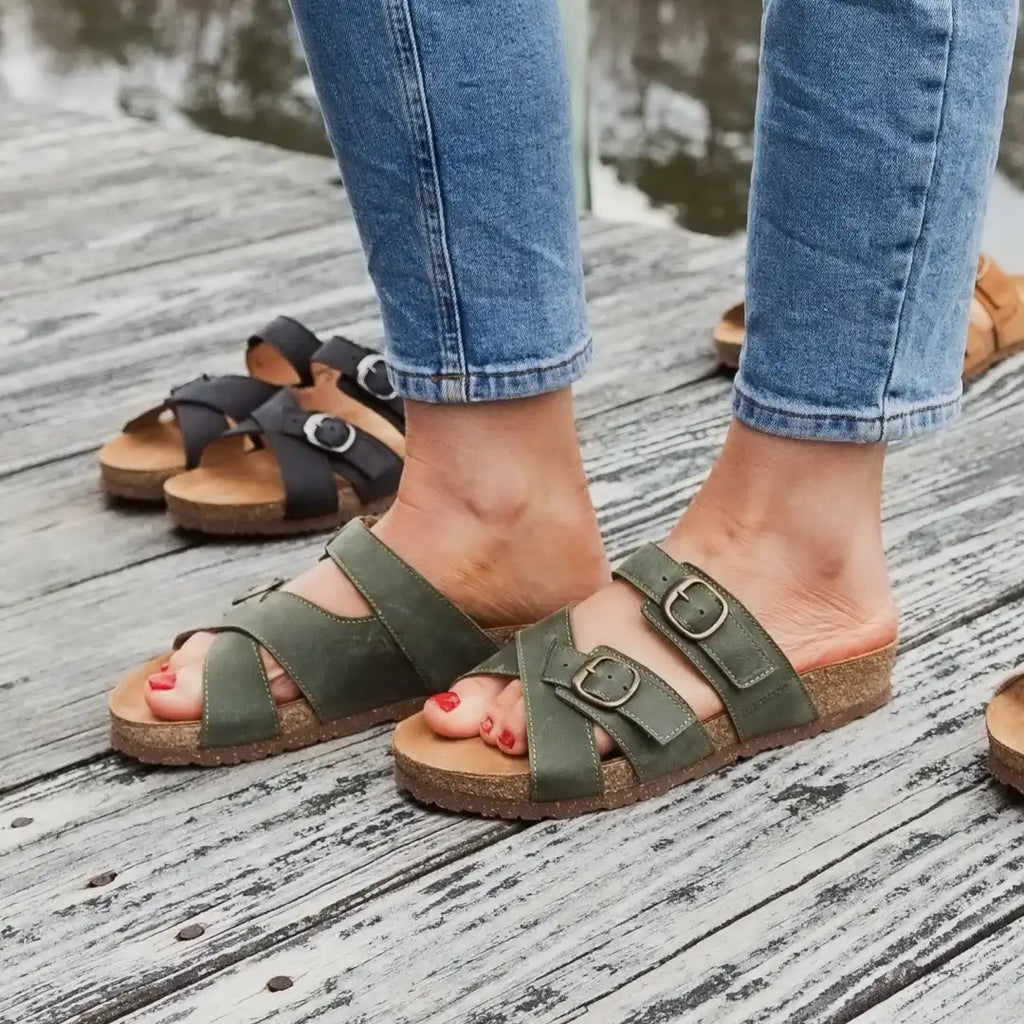 A person wears the Women's Heidi Leather Sandal - Medium in green with adjustable double straps and cork soles on a wooden dock; black and brown pairs are visible in the background.