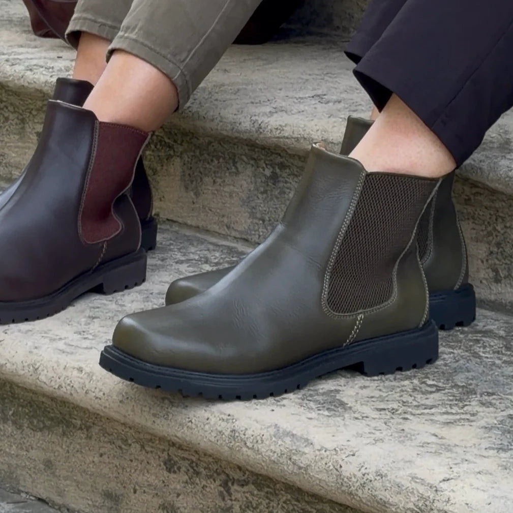 Two people sit on stone steps, each wearing Chelsea boots; one pair is the Women’s Brenner Chelsea Boot in brown water-resistant leather with a dark red elastic side, while the other is a lightweight men’s boot in dark green with a brown mesh side.