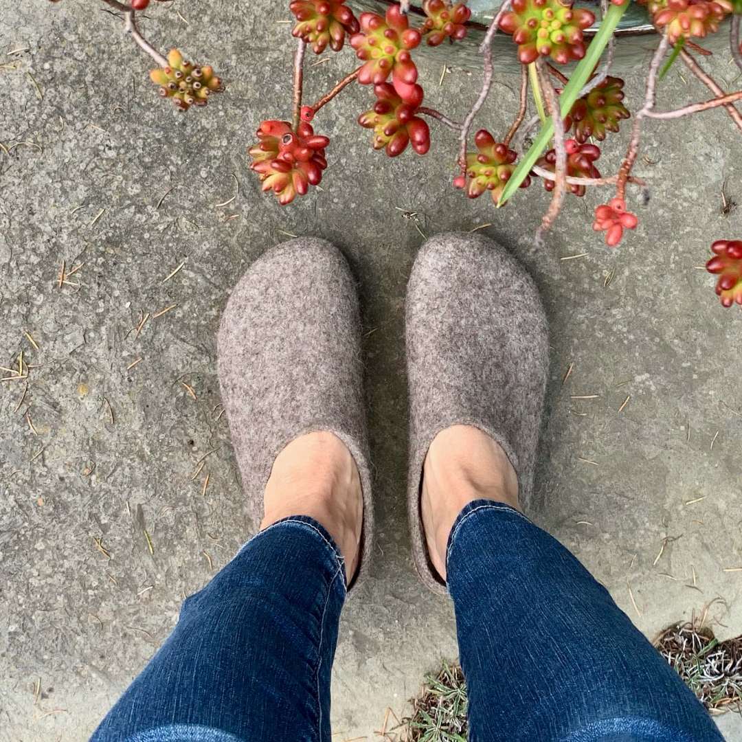 A person in Women's Eco Wool Clogs made from a sustainable wool blend and blue jeans stands on concrete, with red and green succulents visible in the top right corner, adding a natural touch.