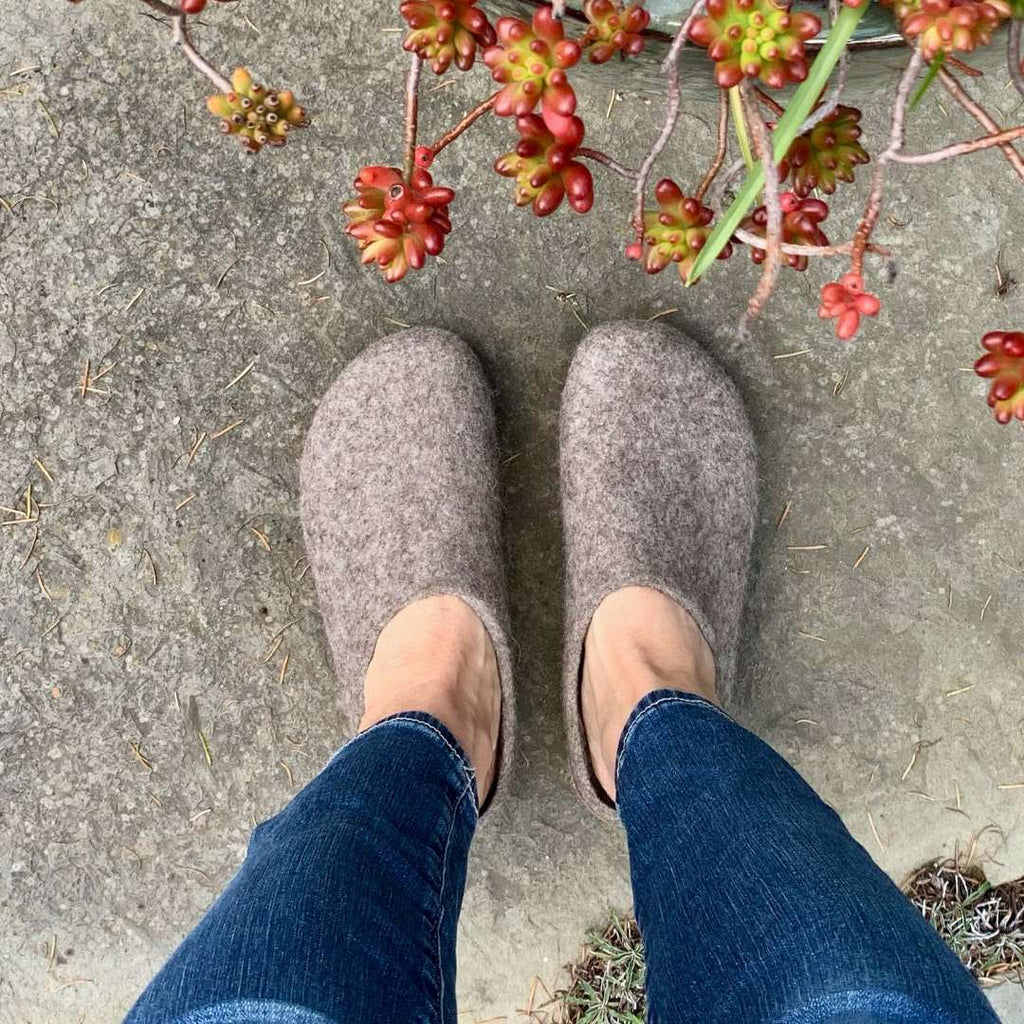 A person in Women's Eco Wool Clogs made from a sustainable wool blend and blue jeans stands on concrete, with red and green succulents visible in the top right corner, adding a natural touch.