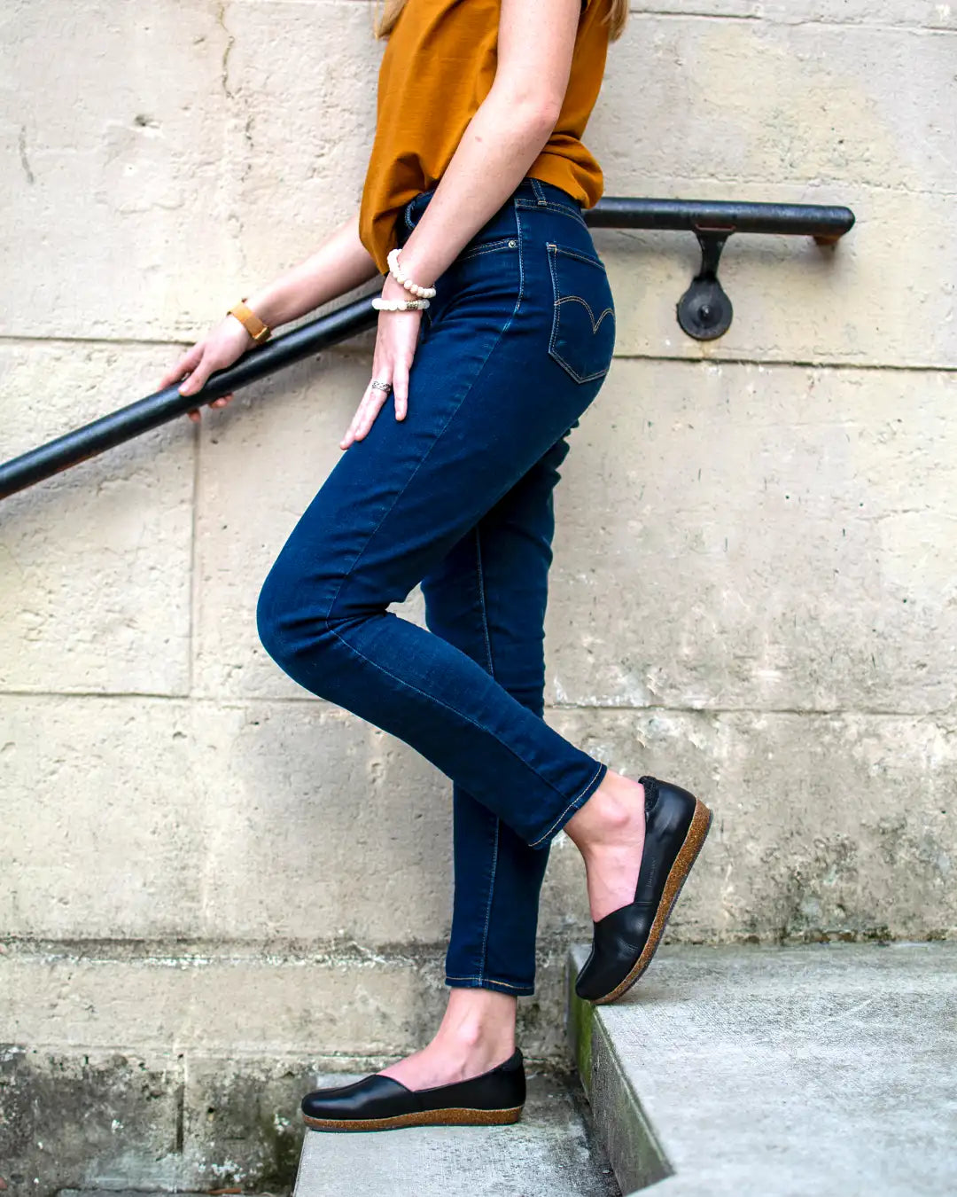 A person stands sideways on stairs holding a black railing, wearing a mustard top, dark blue jeans, and black Women's Liesl Leather Skimmer shoes with anatomical support soles. The person's head is not visible in the image.