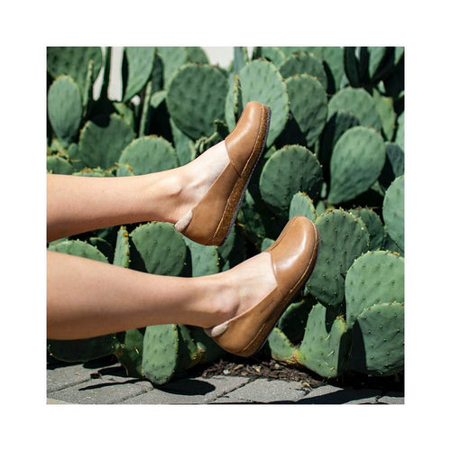 Two legs in Women's Liesl Leather Skimmer shoes, featuring a sleek brown leather upper, are extended against green cactus plants.