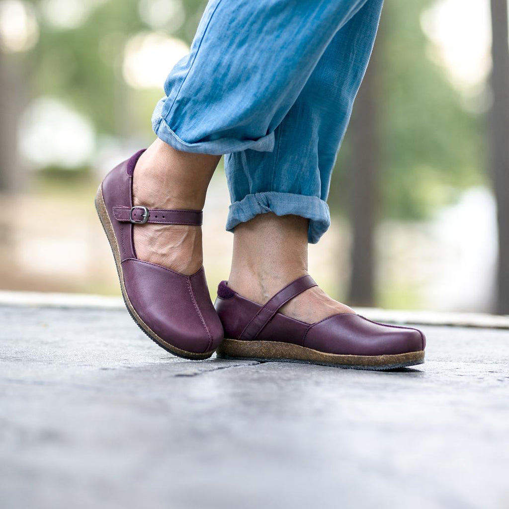 A person stands confidently on a paved surface in Women's Eva Leather Mary-Jane shoes, featuring an adjustable strap, paired with blue rolled-up jeans. The blurry outdoor background adds mystery.