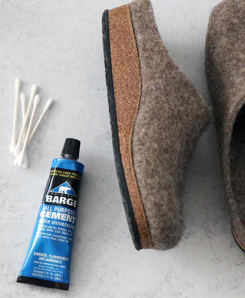 A tube of Barge Shoe Repair Cement, brown felt slippers, and several cotton swabs rest on a gray surface.