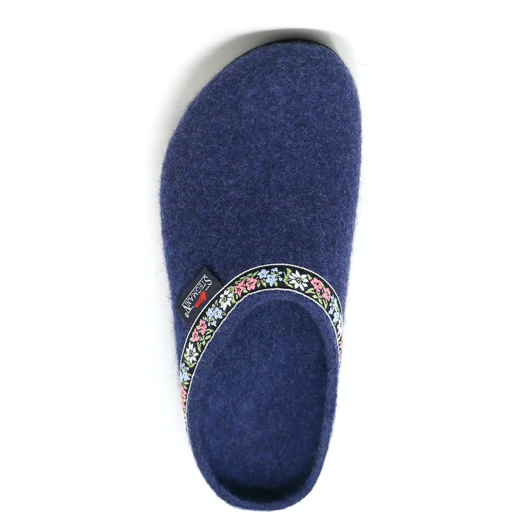 The 'Women's Edelweiss Wool Clog - Medium' is a single blue slipper made of cozy Merino wool, featuring a floral-patterned trim around the opening that resembles hand-embroidered bands.