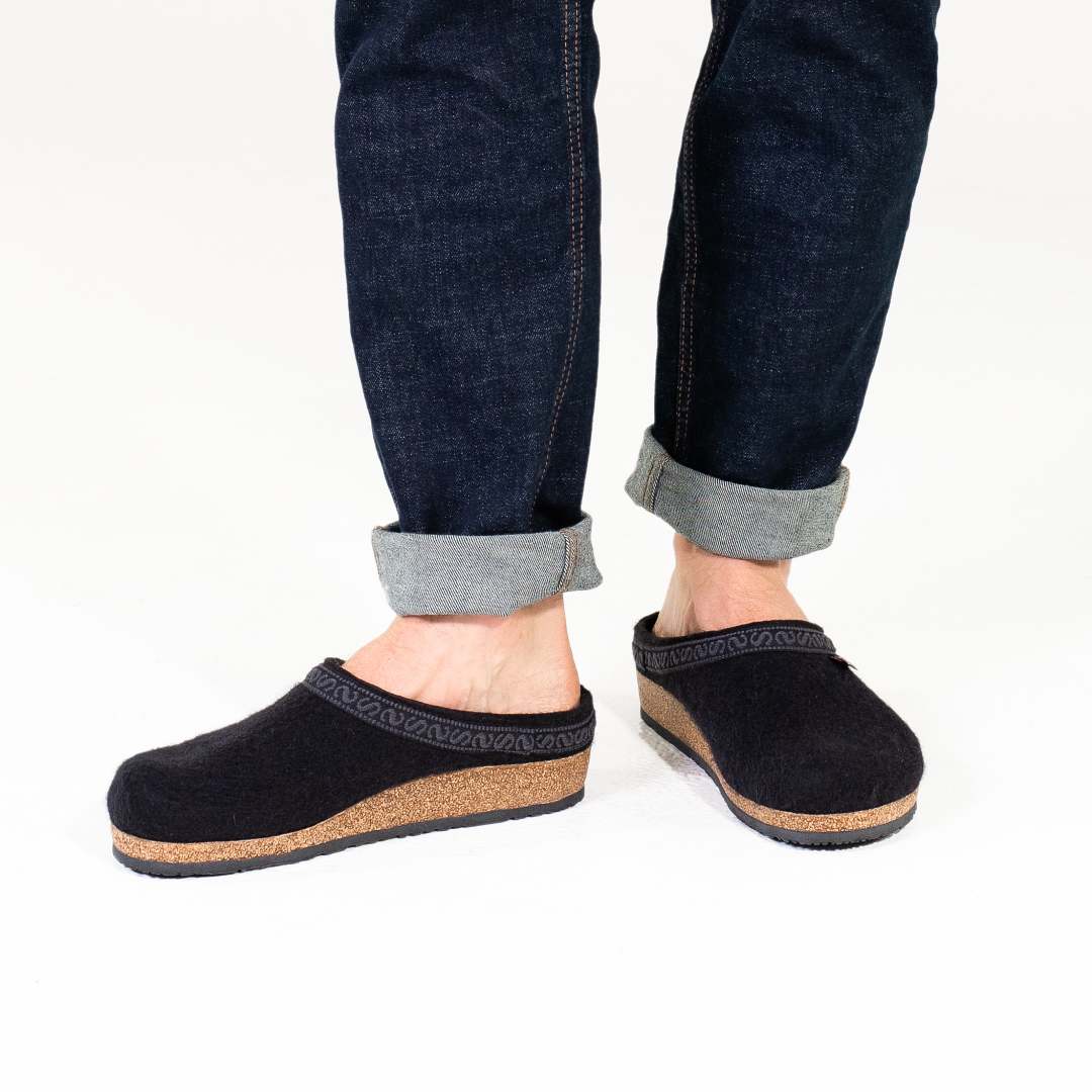 A person sporting rolled-up dark jeans pairs them with the Men's Original 108 Wool Clog, featuring sleek black slip-on design with cork and Merino wool accents for a stylish touch. The addition of a support sole ensures comfort and fashion in equal measure, making this clog an ideal choice for any modern man's shoe collection.